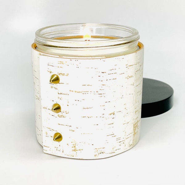 An all natural soy candle is wrapped in a white cork sleeve with 3 gold cone shaped studs