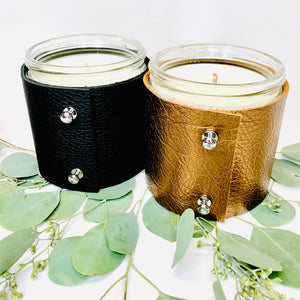 Two 16 oz luxury soy scented candles, one in textured black leather and one in metallic bronze leather with shiny silver button studs