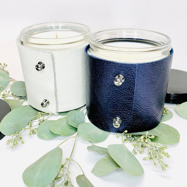 Two 16 oz soy scented candles, one in a fine white leather sleeve and one in s metallic sapphire leather sleeve with two shiny silver button studs