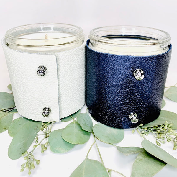 Two 16 oz soy scented candles, one in a fine white leather sleeve and one in s metallic sapphire leather sleeve with two shiny silver button studs